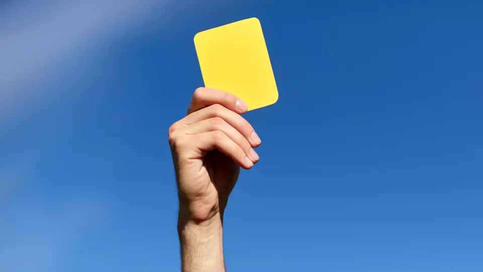 En hand som håller upp ett gult kort mot en blå himmel. Foto.